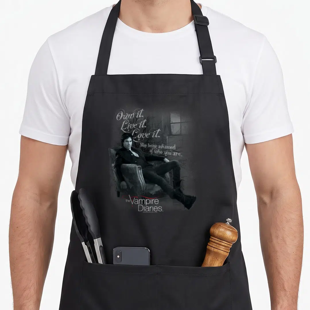 Yourself vampire apron - Man in armchair with script text Own it Live it Love it and Vampire Diaries logo — shown on a black cotton canvas apron against a white background