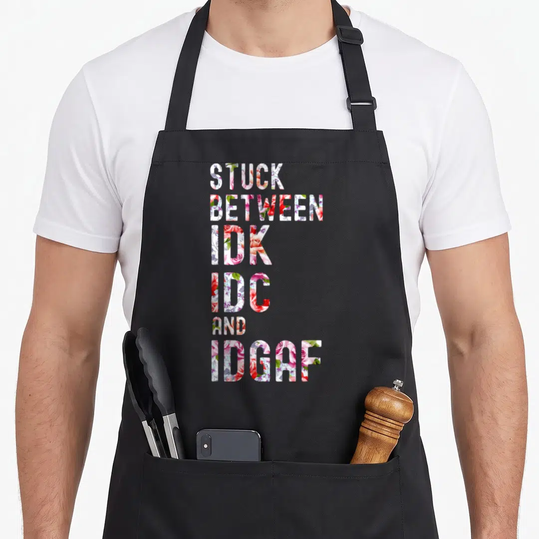 Stuck idgaf apron - Stacked text filled with red and pink floral pattern reading STUCK BETWEEN IDK IDC AND IDGAF — shown on a black cotton canvas apron against a white background