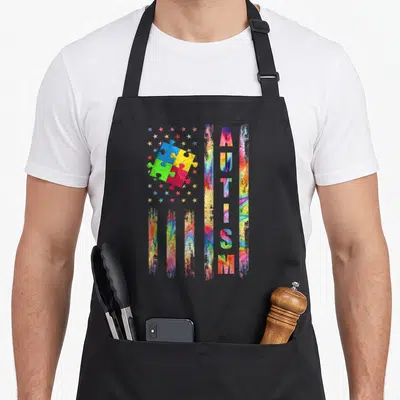 Awareness american flag apron - Distressed flag with four colorful puzzle pieces and vertical AUTISM text on black bib — shown on a black cotton canvas apron against a white background