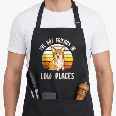 Dog friend corgi aprons - Cartoon corgi sits before striped sun under text I've Got Friends In Low Places — shown on a black cotton canvas apron against a white background
