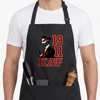 Funny psi alpha apron - Man in suit and mask with 1911 and KAPSI text in red and black — shown on a black cotton canvas apron against a white background