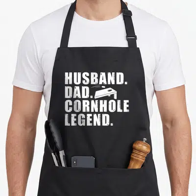 Cornhole husband apron - White distressed text HUSBAND DAD CORNHOLE LEGEND with flying bean bag and board graphic — shown on a black cotton canvas apron against a white background