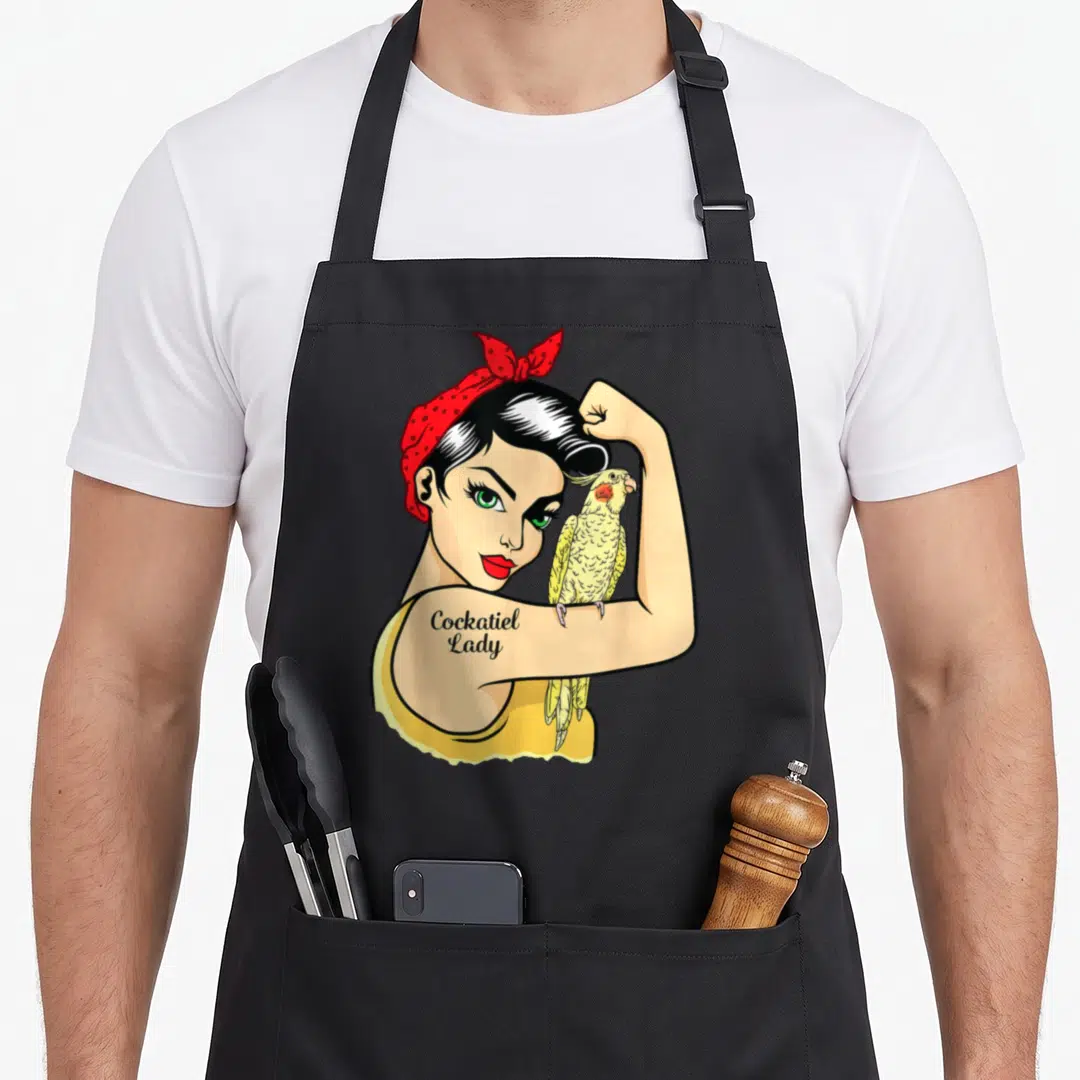 Strong bird apron - Cartoon woman flexing arm with yellow cockatiel perched on bicep and tattoo text — shown on a black cotton canvas apron against a white background