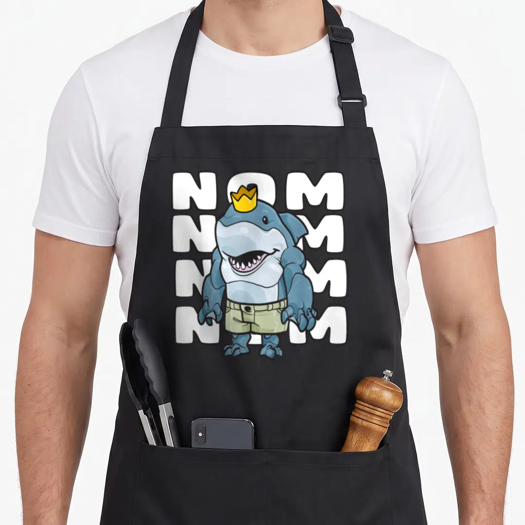 King camp chef apron - Muscular cartoon shark with yellow crown and beige shorts standing in front of four stacked — shown on a black cotton canvas apron against a white background
