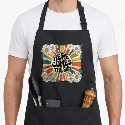 Song come and get it apron - Retro sunburst with HERE COMES THE SUN text surrounded by white daisies and green leaves — shown on a black cotton canvas apron against a white background