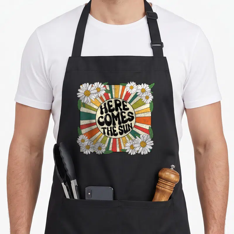Song come and get it apron - Retro sunburst with HERE COMES THE SUN text surrounded by white daisies and green leaves — shown on a black cotton canvas apron against a white background