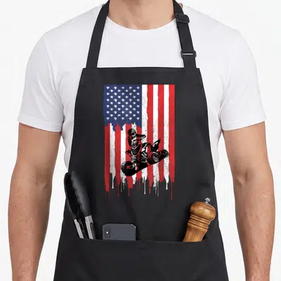 Atv four american flag apron - Black ATV rider jumping over distressed dripping red white blue USA flag design — shown on a black cotton canvas apron against a white background