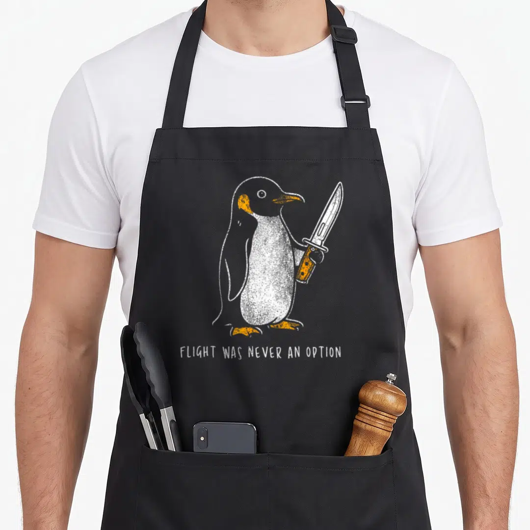 Flight penguin aprons - White and orange line-art penguin holding a knife with text FLIGHT WAS NEVER AN OPTION — shown on a black cotton canvas apron against a white background