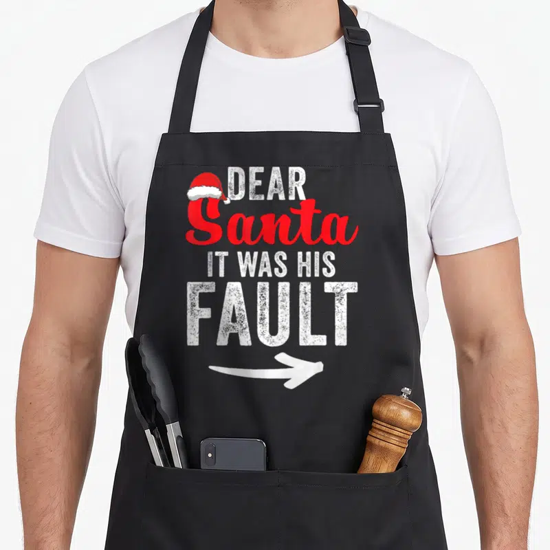 His couples christmas aprons - Stacked white and red text DEAR Santa IT WAS HIS FAULT with a Santa hat and arrow — shown on a black cotton canvas apron against a white background