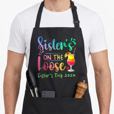 Sister tie-dye apron - Sisters On The Loose gradient text with cocktail, blue and red hearts, and Sister's Trip 2024 — shown on a black cotton canvas apron against a white background
