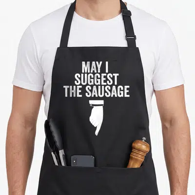 Suggest sausage apron - Distressed white text reading May I Suggest The Sausage above a pointing hand — shown on a black cotton canvas apron against a white background