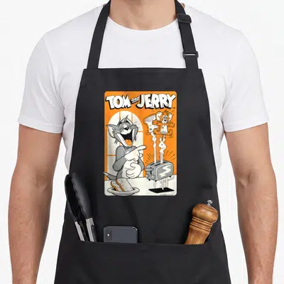 Funny toast apron - Tom pointing at Jerry popping from toaster with toast plate on orange background — shown on a black cotton canvas apron against a white background