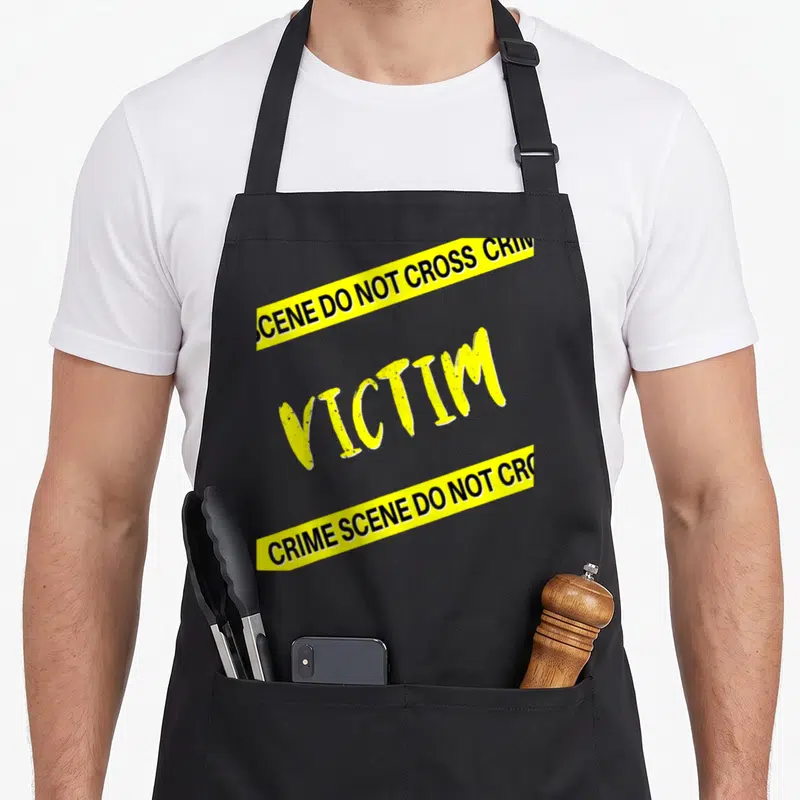 Dinner mystery apron - Yellow VICTIM text between two diagonal crime scene tape strips on black background — shown on a black cotton canvas apron against a white background