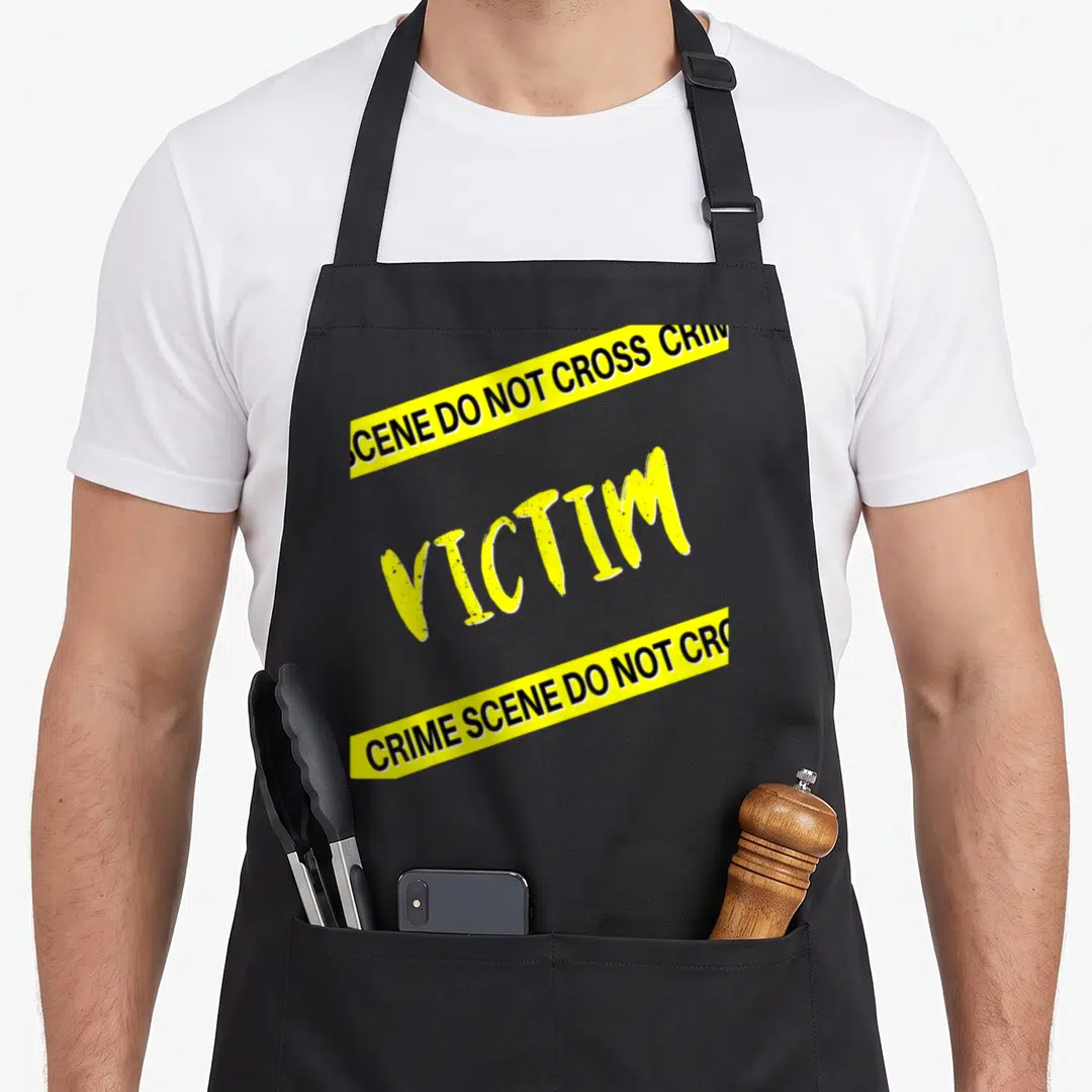 Dinner mystery apron - Yellow VICTIM text between two diagonal crime scene tape strips on black background — shown on a black cotton canvas apron against a white background