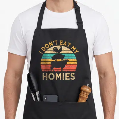 Vegetarian vegan apron - I Don't Eat My Homies text with stacked rooster pig cow silhouettes over striped sun — shown on a black cotton canvas apron against a white background