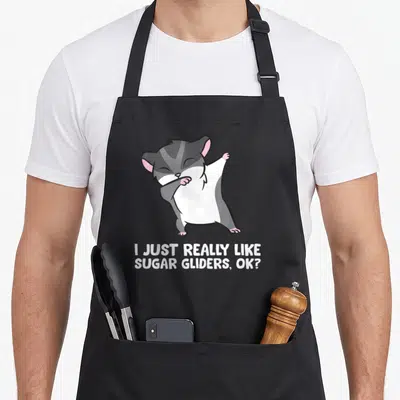 Vintage glider sugar apron - Cartoon sugar glider dabbing above white text I JUST REALLY LIKE SUGAR GLIDERS OK — shown on a black cotton canvas apron against a white background