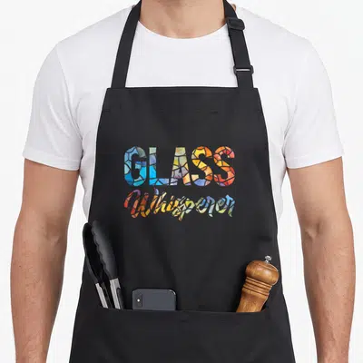 Artist stained apron - GLASS Whisperer text filled with stained glass patterns in blue, yellow, red, green, and purple — shown on a black cotton canvas apron against a white background