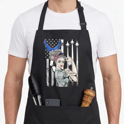 Usaf air force veteran apron - Cartoon woman in camo flexing arm with Veteran tattoo, US flag, three jets, dog tags — shown on a black cotton canvas apron against a white background