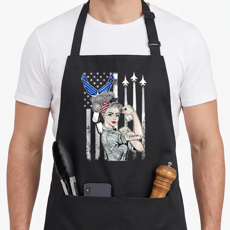Usaf air force veteran apron - Cartoon woman in camo flexing arm with Veteran tattoo, US flag, three jets, dog tags — shown on a black cotton canvas apron against a white background