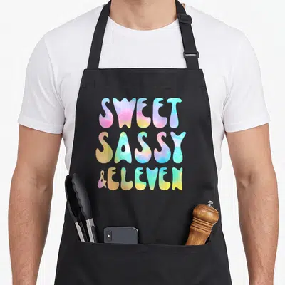 Eleven sassy apron - SWEET SASSY & ELEVEN tie-dye typography in pink blue yellow green purple — shown on a black cotton canvas apron against a white background