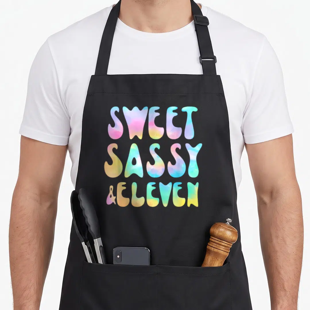 Eleven sassy apron - SWEET SASSY & ELEVEN tie-dye typography in pink blue yellow green purple — shown on a black cotton canvas apron against a white background