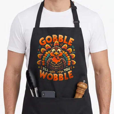 Till thanksgiving apron - Cartoon turkey with orange and blue feathers and text GOBBLE TILL YOU WOBBLE — shown on a black cotton canvas apron against a white background