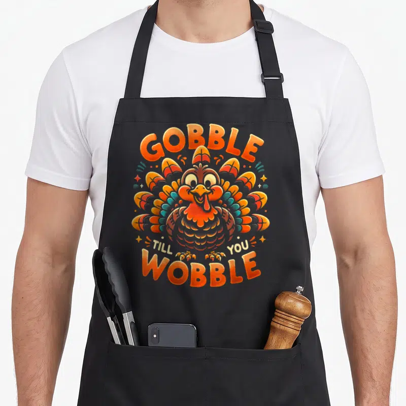 Till thanksgiving apron - Cartoon turkey with orange and blue feathers and text GOBBLE TILL YOU WOBBLE — shown on a black cotton canvas apron against a white background