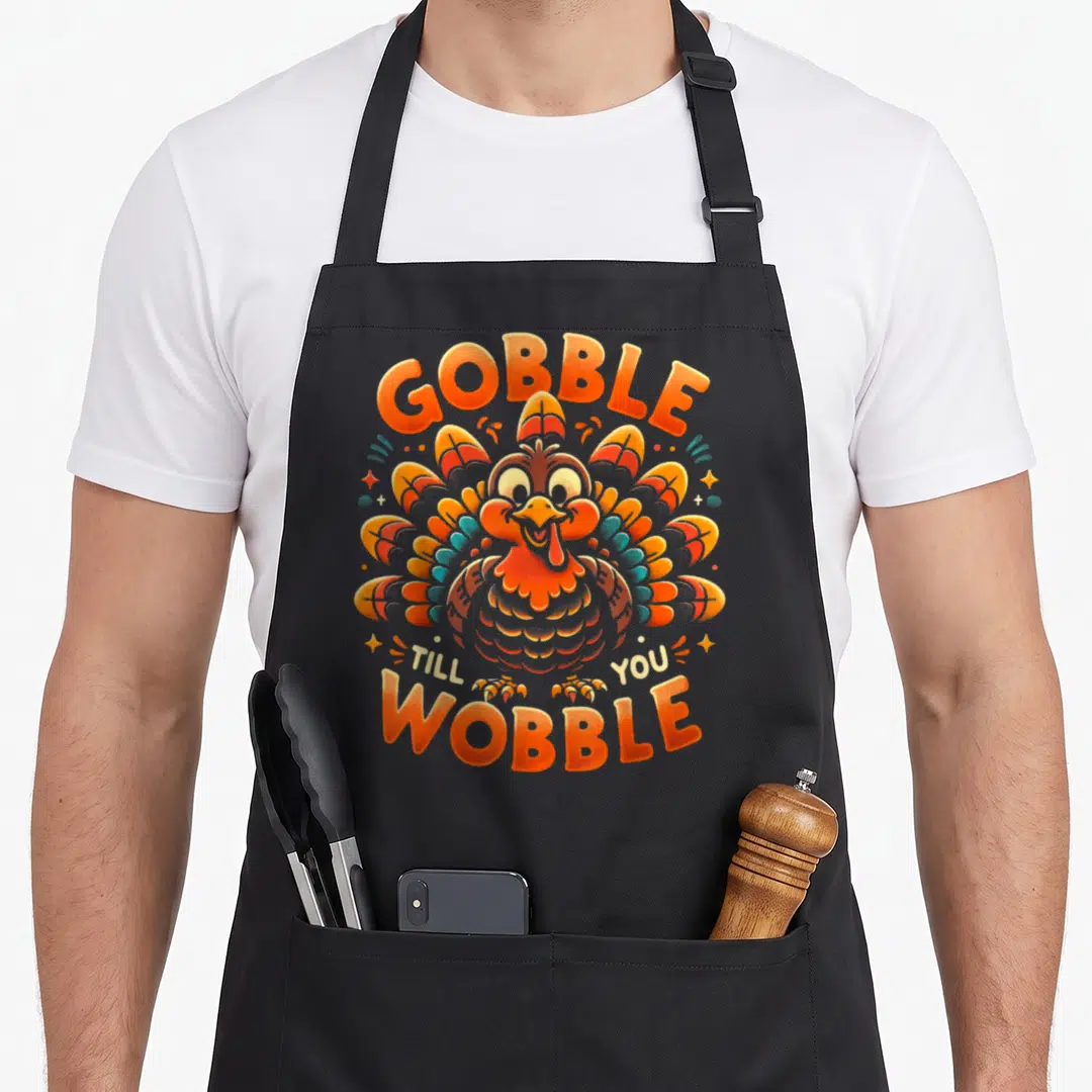 Till thanksgiving apron - Cartoon turkey with orange and blue feathers and text GOBBLE TILL YOU WOBBLE — shown on a black cotton canvas apron against a white background