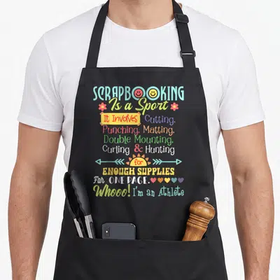 Sport crafting apron - Scrapbooking Is a Sport text design with smiley faces, flowers, hearts, and arrows in multiple — shown on a black cotton canvas apron against a white background
