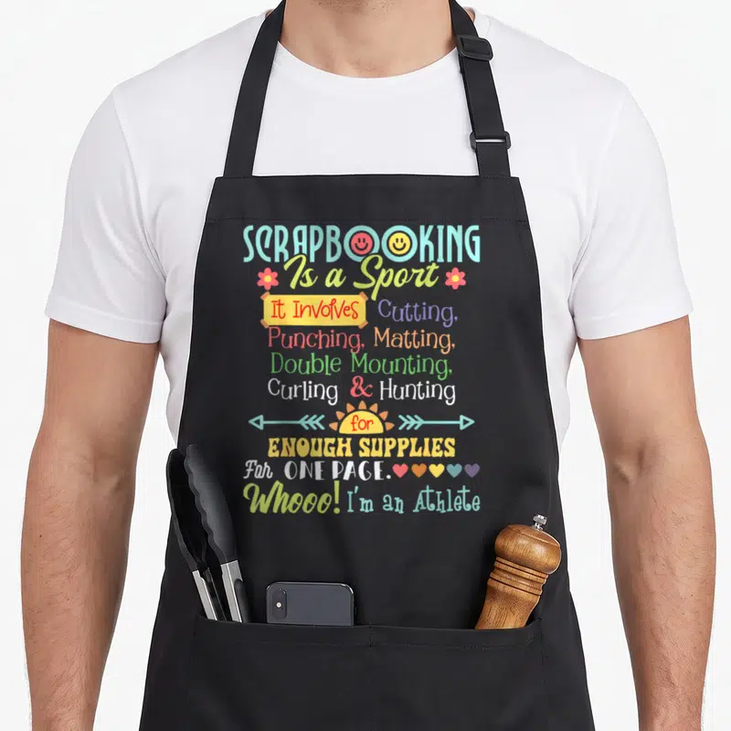 Sport crafting apron - Scrapbooking Is a Sport text design with smiley faces, flowers, hearts, and arrows in multiple — shown on a black cotton canvas apron against a white background