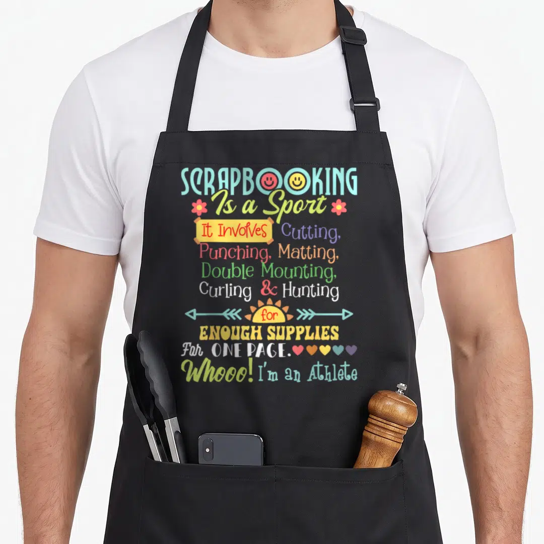 Sport crafting apron - Scrapbooking Is a Sport text design with smiley faces, flowers, hearts, and arrows in multiple — shown on a black cotton canvas apron against a white background