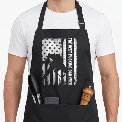 Pucking hockey dad apron - White distressed US flag with hockey player silhouette and vertical text reading THE BEST — shown on a black cotton canvas apron against a white background