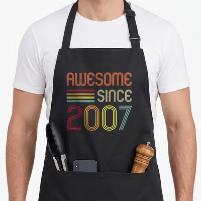 17th birthday apron - Retro stacked text AWESOME SINCE 2007 with orange red yellow and blue-grey stripes — shown on a black cotton canvas apron against a white background