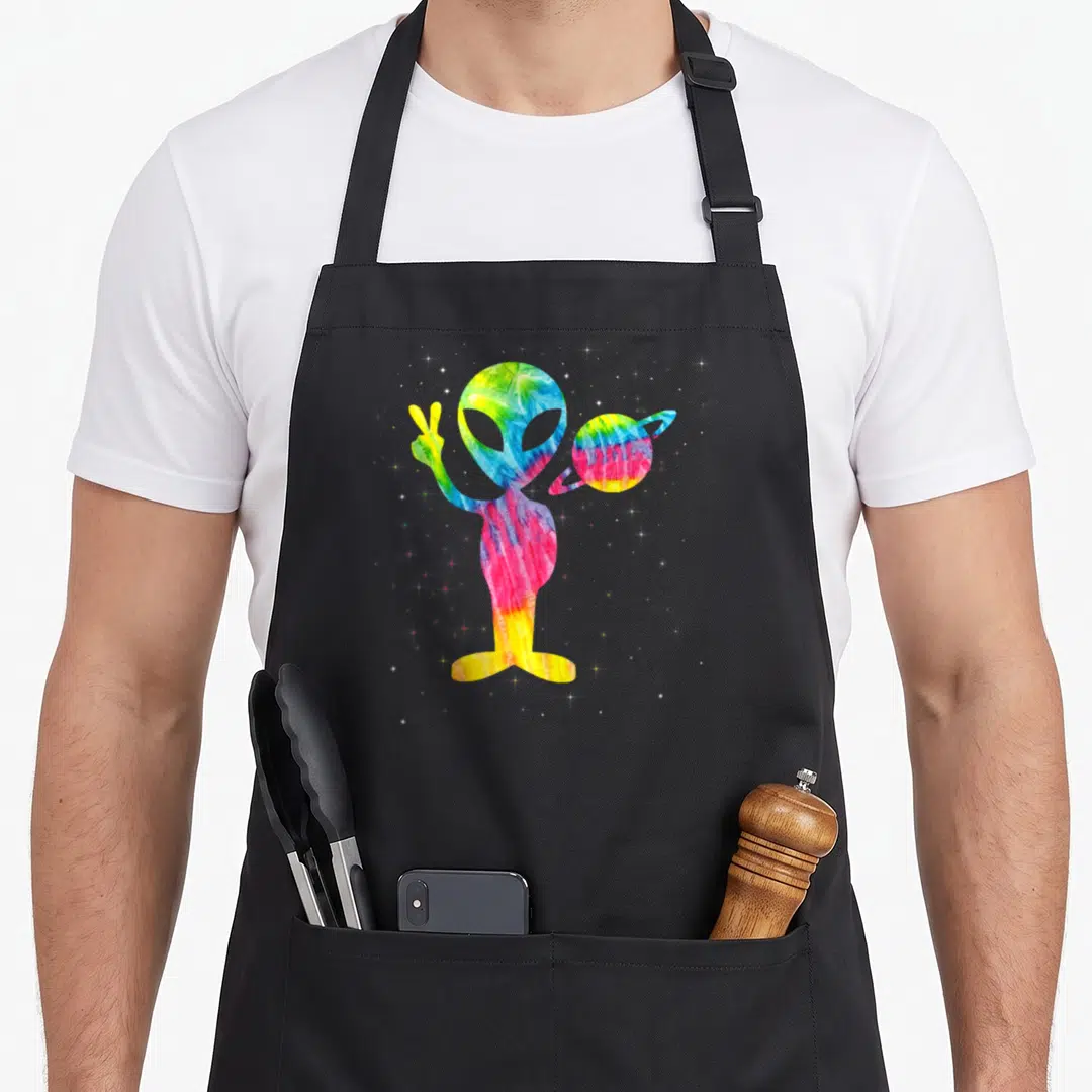 Alien tie-dye apron - Tie-dye alien holding a ringed planet and making a peace sign among scattered stars — shown on a black cotton canvas apron against a white background