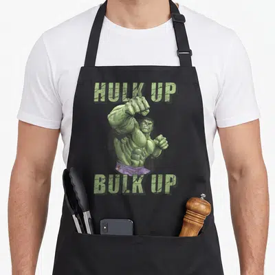Incredible worlds greatest apron - Green muscular figure in purple shorts with HULK UP BULK UP text — shown on a black cotton canvas apron against a white background