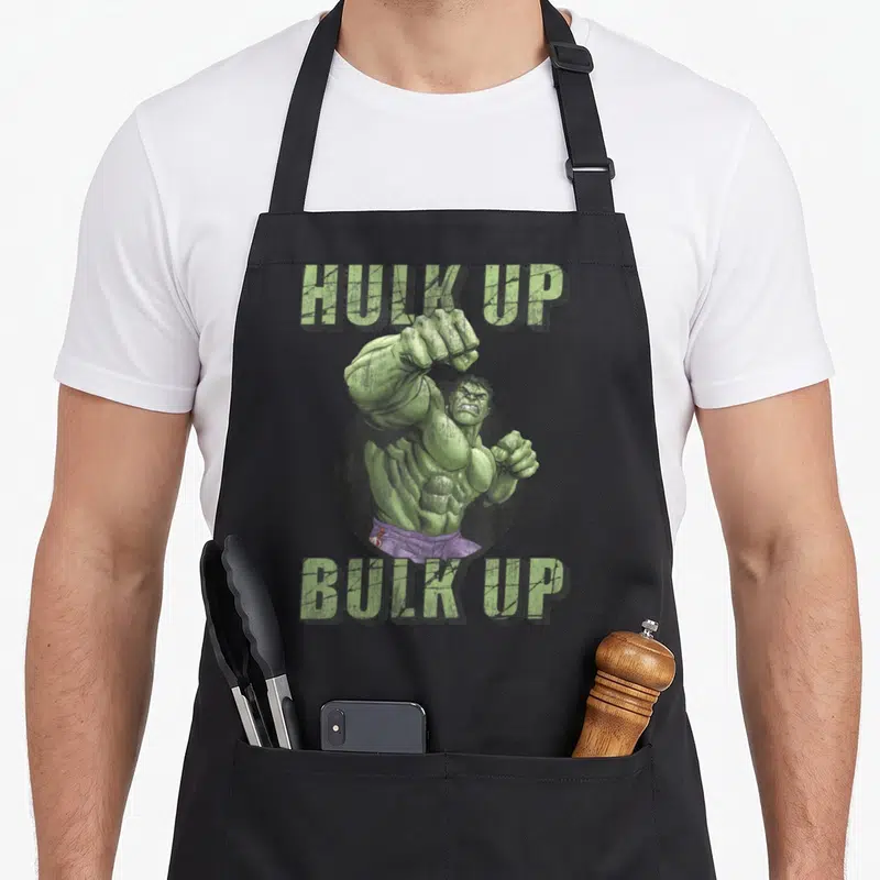 Incredible worlds greatest apron - Green muscular figure in purple shorts with HULK UP BULK UP text — shown on a black cotton canvas apron against a white background