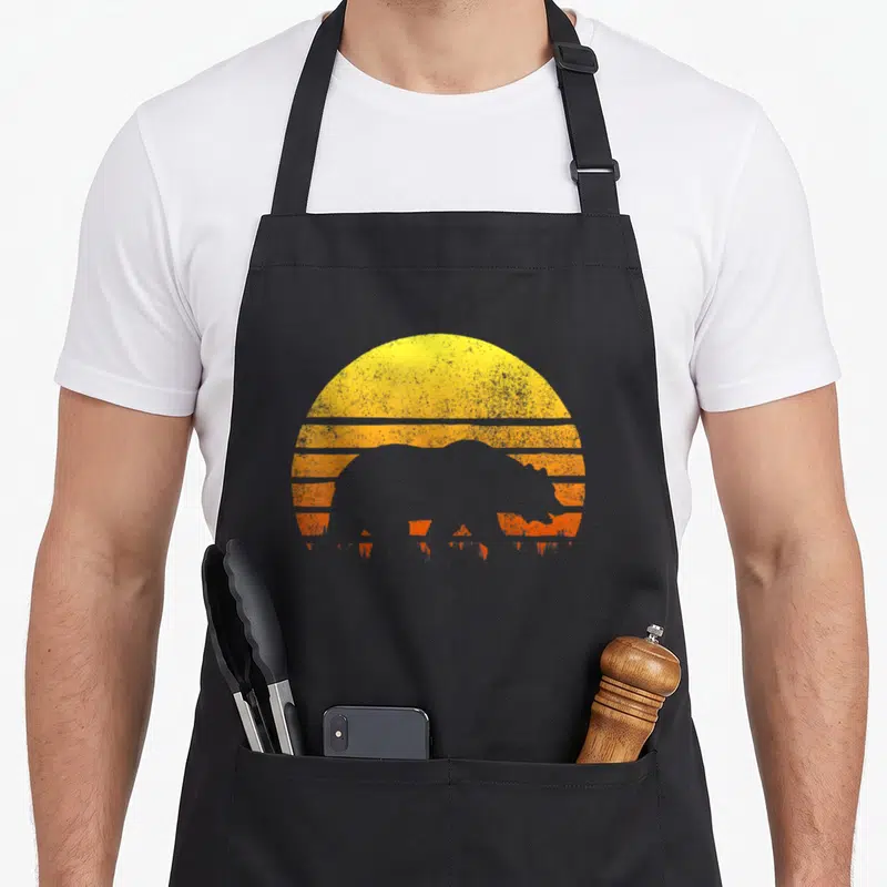Forest bear apron - Retro bison silhouette against yellow and orange striped semi-circle sun — shown on a black cotton canvas apron against a white background