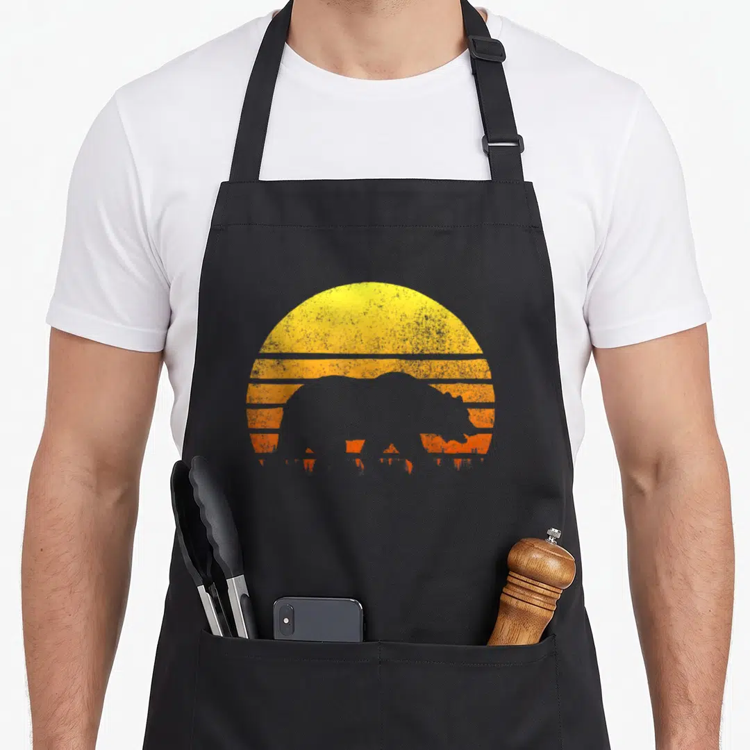 Forest bear apron - Retro bison silhouette against yellow and orange striped semi-circle sun — shown on a black cotton canvas apron against a white background