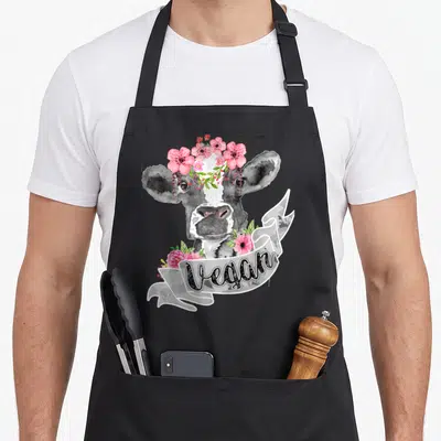 Cow vegan apron - Watercolor cow head with pink flower crown and ribbon banner reading Vegan in black script — shown on a black cotton canvas apron against a white background