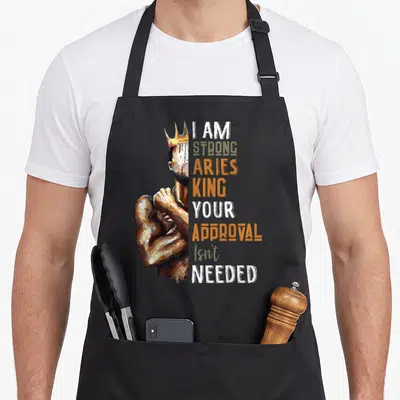 Ary king apron - Painted crowned man flexing arm next to stacked text I Am Strong Aries King Your Approval Isn't Needed — shown on a black cotton canvas apron against a white background