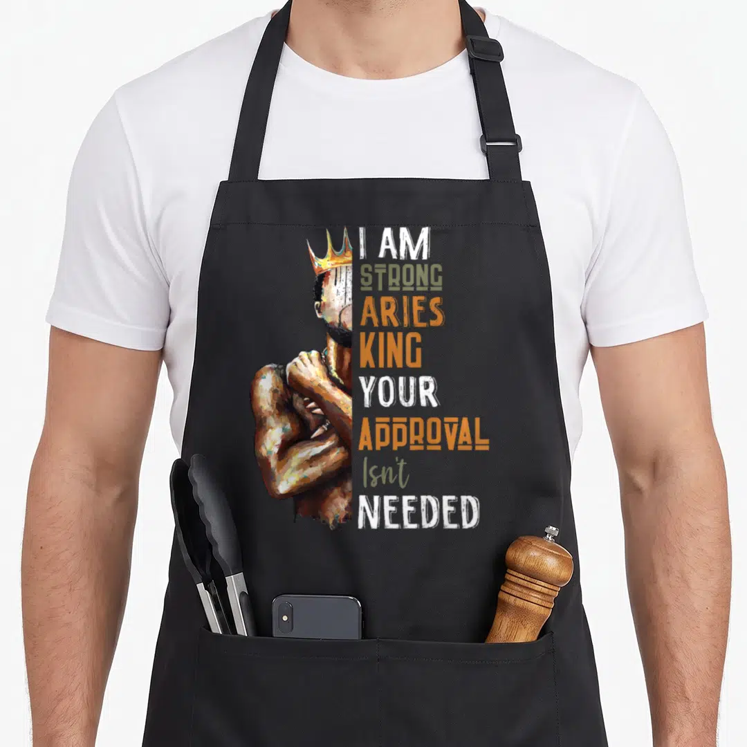 Ary king apron - Painted crowned man flexing arm next to stacked text I Am Strong Aries King Your Approval Isn't Needed — shown on a black cotton canvas apron against a white background