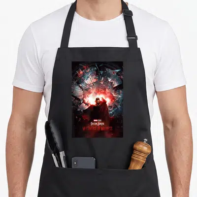 Madness doctor apron - Doctor Strange centered in red blue and black shattered glass with stacked movie title text below — shown on a black cotton canvas apron against a white background