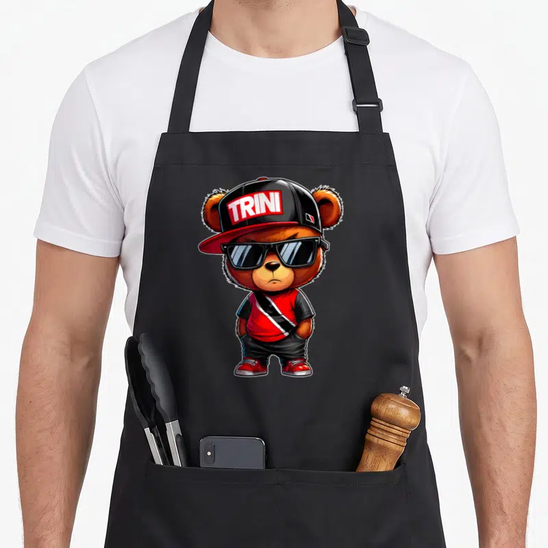 Trini teddy bear apron - Cartoon bear wearing sunglasses and cap with TRINI text in red black and white — shown on a black cotton canvas apron against a white background