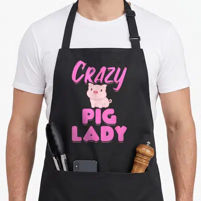 Funny pig lady apron - Pink cartoon pig with arched CRAZY PIG LADY text stacked vertically — shown on a black cotton canvas apron against a white background