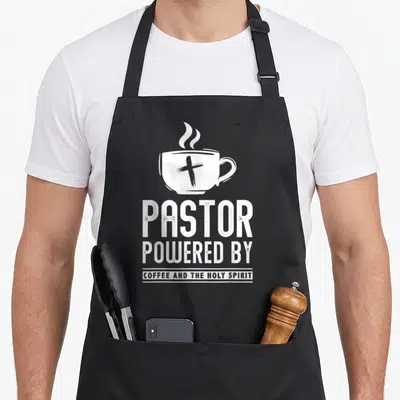Pastor coffee apron - White distressed coffee cup with cross above stacked text pastor powered by coffee and the holy — shown on a black cotton canvas apron against a white background