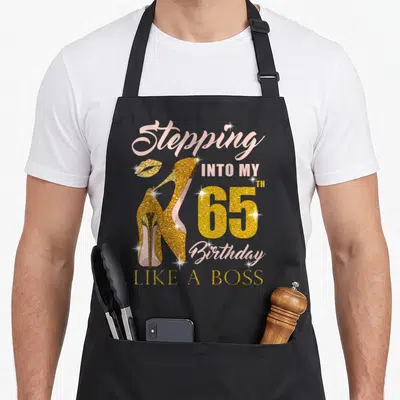 65th bbq boss apron - Gold high heels and lips with Stepping Into My 65th Birthday Like A Boss text — shown on a black cotton canvas apron against a white background