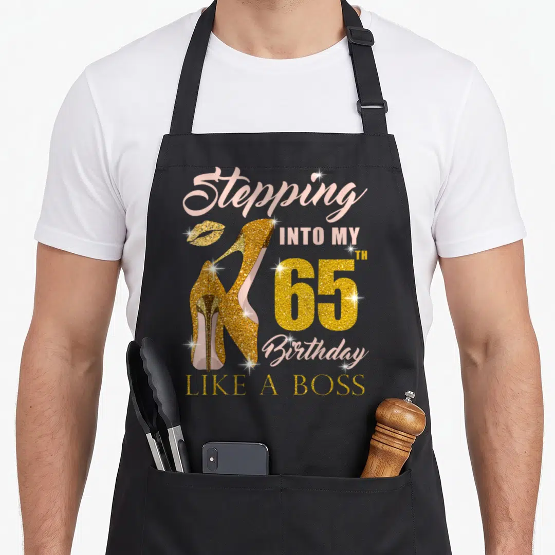 65th bbq boss apron - Gold high heels and lips with Stepping Into My 65th Birthday Like A Boss text — shown on a black cotton canvas apron against a white background