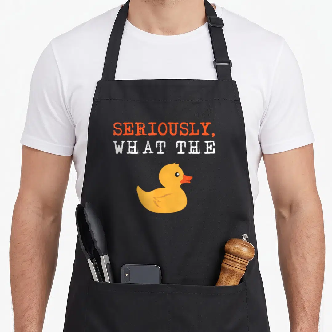 Seriously rubber apron - Orange and white text reading SERIOUSLY WHAT THE above a yellow cartoon duck — shown on a black cotton canvas apron against a white background