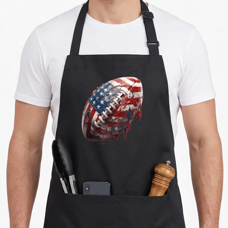 Flag football apron - Realistic football with American flag pattern and red white blue paint dripping — shown on a black cotton canvas apron against a white background