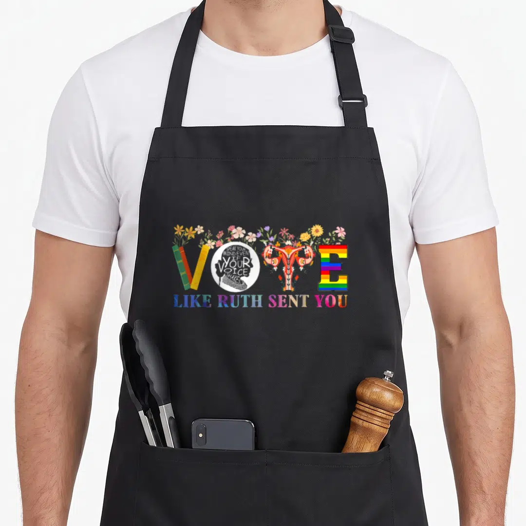 Ruth novelty apron - VOTE formed by books uterus rainbow E flowers with SPEAK YOUR MIND text — shown on a black cotton canvas apron against a white background